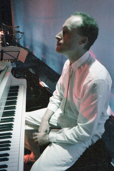 Boris Conley Handspan Theatre man sitting at a piano with hands in his lap wearing white shirt and trousers backlit in blue and pink lighting