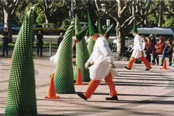 People in white coats, red trousers and tall cone-shaped black hats, free-standing 2-metre green foam cones and standard orange plastic roadwork cones