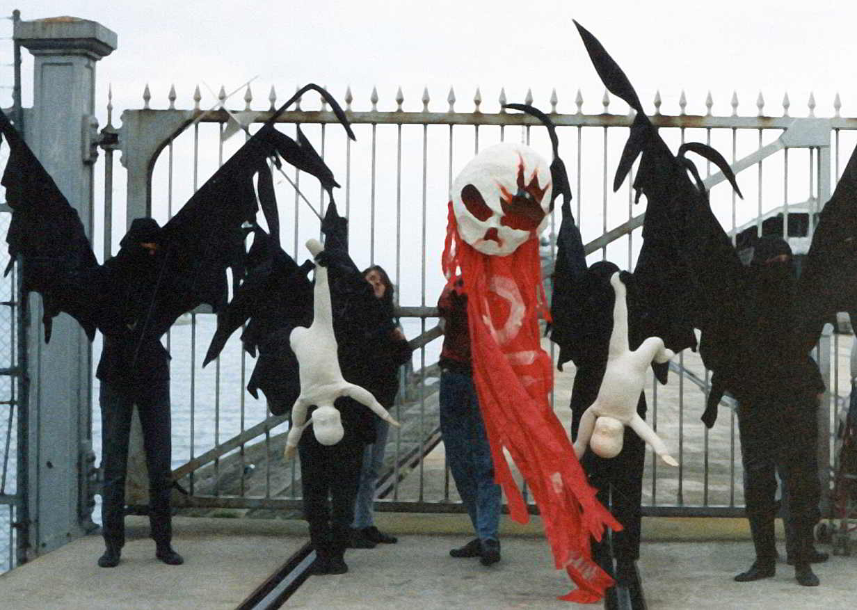 Handspan Theatre Protests skeleton head above red flag flanked on each side by a white baby hanging upside down and a black-winged figure