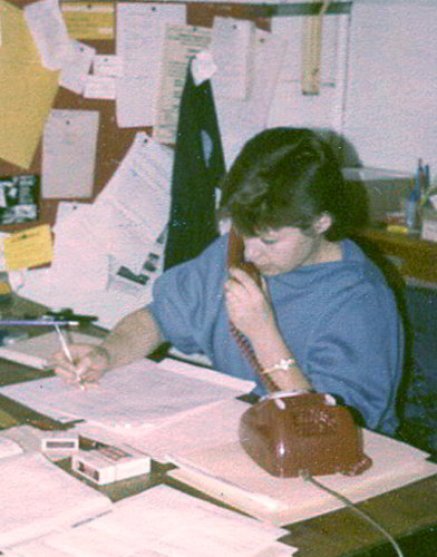 Diana Kidd Handspan Theatre colour picture of young woman with pencil in hand and telephone at her ear seated at a desk covered in papers