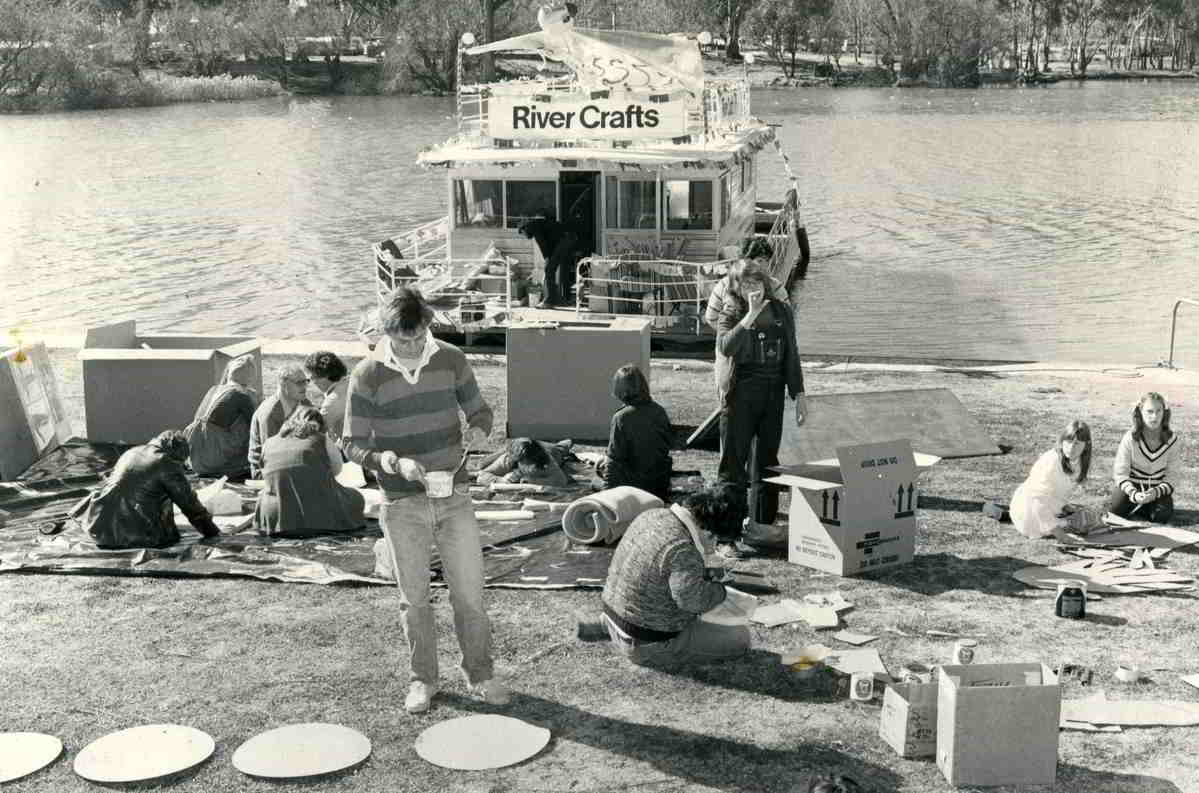 Rivercraft, Mildura John Rogers with craft materials on the bank of a river with houseboat on the water