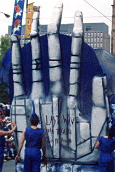 several people in blue with blue wave-like headwear guide a giant blue hand in city street parade